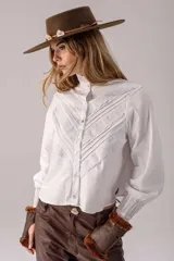 Camisa blanca de algodón con cuello alto, mangas largas abullonadas y cierre frontal de botones. Presenta un diseño delantero con alforzas y detalles de bordado inglés en forma de V.