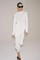 Jersey de punto liso blanco, con cuello redondo y mangas largas. Presenta un diseño de bajo asimétrico pronunciado.