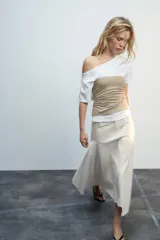 Falda midi de tiro medio color gris, con diseño asimétrico en el bajo, bolsillos frontales y traseros, y cierre frontal con cremallera y botón.