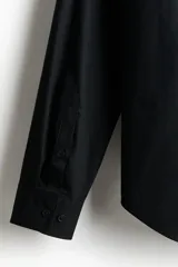 Camisa negra de algodón con cuello italiano, tapeta clásica y canesú en la espalda. Mangas largas con tapeta y botón, y cierre ajustable de botón en los puños. Bajo levemente redondeado. Corte ajustado que realza el contorno del cuerpo creando una silueta entallada.