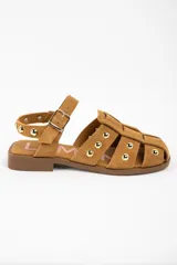 Sandalia tipo cangrejera de gamuza color camel, con diseño de tiras entrelazadas y tachas metálicas doradas. Cuenta con cierre de pulsera con hebilla ajustable y suela plana de goma.