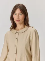 Camisa de algodón color beige con cuello bebé, cierre frontal con botones y cortes curvos que estilizan la figura.