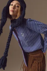 Camisa de algodón a rayas verticales azules y blancas, con corte holgado, cuello clásico y detalle de bordado de estrellas en el pecho. Presenta terminaciones en contratono rojo en la cartera frontal.