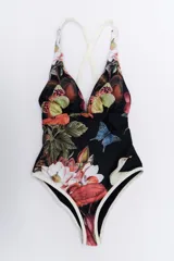 Malla enteriza con estampado botánico y de mariposas sobre fondo negro, con escote en V profundo y breteles cruzados en la espalda. Presenta detalles de ribetes en color crema en los bordes.