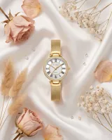 Reloj de pulsera analógico con caja redonda de metal dorado y bisel con incrustaciones de cristales. Esfera blanca con números romanos y malla de acero tipo milanesa en tono dorado.