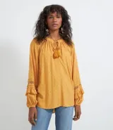 Blusa de manga larga color mostaza con textura de puntos en relieve (dobby). Presenta cuello redondo con abertura en V y cordón ajustable con borla (tassel) en el escote. Las mangas tienen detalles de entretejido calado (entremedios) y volados con terminación fruncida en los puños.