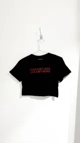 Camiseta negra de corte cropped con estampa frontal en color rojo que dice "Fear a bit less, live a bit more".