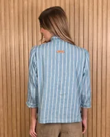 Camisa de algodón color blanco con rayas verticales rosadas, cuello alto con volados, mangas tres cuartos abullonadas con puños elastizados con volados y cierre frontal de botones oscuros.