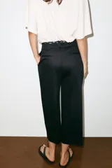 Pantalón de popelina de tiro alto y pernera ancha, de color negro. Presenta pinzas delanteras, bolsillos laterales y falsos bolsillos de vivo en la espalda. Incluye un cinturón combinado tipo pañuelo trenzado con tiras colgantes y cuentas de madera. Cierre frontal con cremallera, botón interior y gancho.