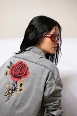 Camisa de jean liviano en tono celeste claro, con corte oversize, cuello clásico, cierre frontal con botones metálicos, bolsillo en el pecho y detalle de bordado en el puño.