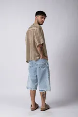 Camisa de manga corta con cuello solapa, corte holgado y cierre frontal de botones. Presenta un diseño de rayas verticales en tonos beige, negro y blanco.