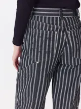 Pantalón de tiro medio y pierna ancha confeccionado en denim de algodón grueso. Presenta un diseño a rayas verticales en tonos azul y celeste, con costuras en contraste color rojo, bolsillos laterales tipo cargo y remaches metálicos.