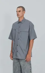 Camisa de manga corta de poplin, color gris, con silueta oversize. Presenta dos bolsillos delanteros, uno con solapa y otro con ojal y botón.