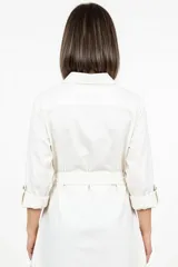 Sobre camisa de corte largo y estilo minimalista, color blanco, con cuello camisero, cierre frontal de botones y cinturón ajustable a la cintura. Presenta mangas largas con puños y bolsillos laterales.