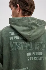 Canguro oversize de algodón color verde militar con efecto lavado. Cuenta con capucha y diseño con estampas tipográficas y texto bordado en la parte trasera.