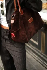 Bolso tipo duffle color verde militar, confeccionado en gamuza, con manijas de cuero marrón. Incluye bolsillos laterales, uno con clip, cierre posterior y un bolsillo interno amplio. Detalle de bandera y logo bordado en los laterales.