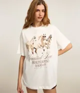 Remera oversized de algodón color crudo, con cuello redondo y mangas cortas. Estampa trasera con texto "Amor Peligroso" y listado de ciudades con fechas, en tipografía estilo western.