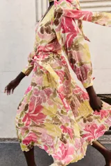 Vestido midi con estampado floral en tonos rosados, verdes y ocres. Presenta cuello con lazo, escote en pico, mangas largas con puños y cintura elástica con cinturón ajustable del mismo tejido. La falda cuenta con un diseño plisado y forro interior.