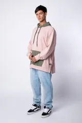 Buzo tipo canguro con capucha, confeccionado en una combinación de algodón rústico y microfibra. Presenta un diseño bicolor en tonos rosado y verde militar, con bolsillo frontal tipo canguro en contraste, cordones ajustables y estampa personalizada en el frente.