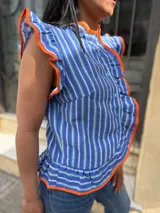 Blusa sin mangas a rayas azules, blancas y verde agua, con cuello alto y cierre frontal de botones. Destaca por sus volados ribeteados en color naranja en el cuello, hombros y la botonera.