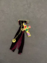 Pin decorativo con forma de cruz dorada de estilo vintage, adornada con piedras incrustadas. La pieza incluye una base de tela de terciopelo desflecada, una cadena metálica y un pequeño dije colgante.
