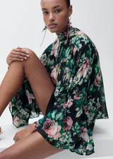 Blusa de gasa con estampado floral en tonos rosa, verde y negro. Tiene cuello alto, mangas largas con volantes y cierre de lazo en la parte posterior.
