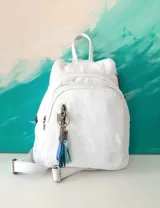 Mochila de cuero blanca con herrajes de metal envejecido y cierres YKK. Cuenta con un bolsillo frontal con cierre y llavero decorativo con borlas en tonos azules.