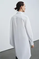 Camisa de popelín de algodón con cuello solapa, manga larga con puños y bajo asimétrico. Presenta cierre frontal con botones ocultos.