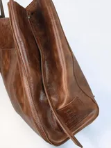 Bolso tipo tote grande de cuero genuino marrón, con asas de hombro dobles, bolsillo exterior con botón a presión y llavero colgante con la marca grabada.