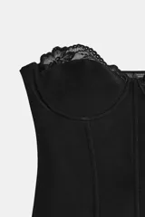 Corsé de escote corazón y hombros descubiertos, confeccionado en tejido elástico color negro. Presenta copas con aros y varillas interiores, con detalle de encaje tono sobre tono en el borde superior.