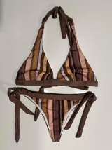 Conjunto de bikini con estampado a rayas verticales en tonos marrones, beige y blanco. El corpiño es de corte triangular con tirantes que se atan al cuello y a la espalda. La bombacha es de corte clásico con tiras laterales para ajustar.