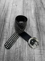 Cinto negro de cuero sintético con tachas plateadas y hebilla redonda plateada con detalles labrados.