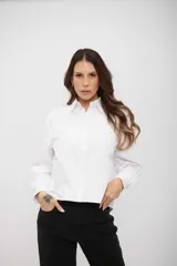 Camisa blanca de corte recto y largo cropped, con cuello clásico, cierre frontal de botones y mangas largas con puños abotonados. Presenta un sutil estampado de pequeños puntos distribuidos por toda la prenda.
