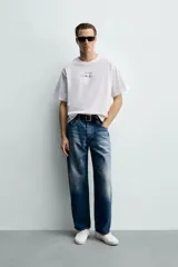 Camiseta de corte boxy fit, color blanco, con cuello redondo y manga corta. Presenta una aplicación de etiquetas decorativas a contraste en el centro del pecho.