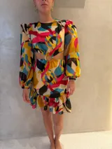 Vestido corto de manga larga abullonada con aberturas laterales en la cintura y detalle de argolla central. Presenta un estampado colorido con motivos abstractos y un ruedo con volado.