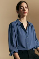 Camisa de manga larga color azul, con cuello clásico y cierre frontal de botones. Presenta un diseño fluido con detalles de vainilla en los hombros y puños elásticos con volados.