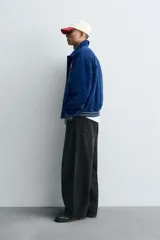 Cazadora estilo varsity confeccionada en pana de algodón azul, con interior ligeramente acolchado. Presenta cuello solapa, cierre frontal con botones a presión, bolsillos laterales de vivo y acabados en rib a rayas en puños y cintura. Incluye múltiples parches bordados decorativos en el frente.