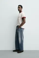 Camiseta blanca de corte relajado (relaxed fit) con cuello redondo y manga corta. Presenta un bolsillo plastrón en el pecho y un estampado de texto en color marrón rojizo en la espalda que dice "SOMETIMES SÍ, SOMETIMES BYE" (reemplazando la 'O' de 'SOMETIMES' por un corazón).