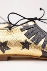 Zapatos tipo blucher artesanales de napa dorada con estampado de estrellas negras. Presentan un adorno de flecos de napa negra en el empeine, cierre con cordones y suela de cuero.