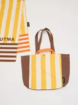 Bolso tipo tote con diseño de rayas verticales en amarillo y blanco, con laterales, base y correas en color marrón. Incluye asas cortas en color naranja y cierre con botón a presión.