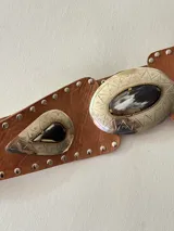 Cinto tipo faja de cuero color beige, con apliques metálicos plateados de forma ovalada y de gota, decorados con piedras oscuras y tachas metálicas a lo largo de toda la pieza.