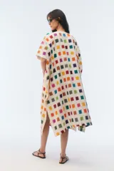 Kaftan amplio de largo midi y escote redondo, con mangas cortas caídas, aberturas laterales y ruedo asimétrico en puntas. Confeccionado en algodón, presenta un estampado de pequeños cuadrados multicolores sobre base blanca.