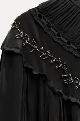 Vestido corto negro con cuello redondo, mangas largas abullonadas con puños elásticos y detalle de encaje y bordados con abalorios. Incluye lazo para ajustar en la cintura.