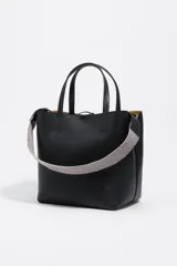 Bolso de hombro tipo tote bag de piel vacuna color beige, con doble asa de mano y correa ajustable de tejido. Presenta un cierre de cordón en la parte superior y un logo metálico en relieve en el frente. Incluye bolsillo interior con cierre de cremallera.