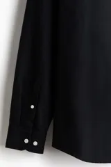 Camisa de algodón estilo Oxford, color negro, con cuello americano abotonado, bolsillo en el pecho y cierre frontal con botones blancos.