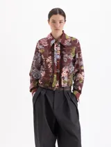 Blusa de manga larga con estampado floral y geométrico en tonos marrones, con cuello clásico, cierre frontal con ganchos metálicos y detalle de volados laterales en la tapeta.