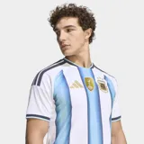Camiseta de fútbol Adidas de la selección argentina, modelo Primera Equipación 26. Diseño a rayas verticales blancas y celestes con un efecto degradado en las franjas celestes. Presenta cuello redondo con ribete azul oscuro y tres franjas de Adidas en los hombros en azul oscuro. El escudo de la AFA y el logo de Adidas son de color dorado.