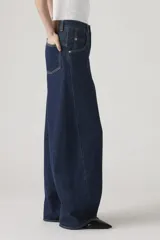 Jean de corte baggy y silueta barrel, confeccionado en denim rígido de color azul oscuro. Presenta costuras laterales curvadas que definen su forma característica, tiro alto y cierre frontal con botón y cremallera.