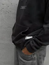 Canguro negro de corte holgado con capucha ajustable mediante cordones, bolsillo frontal tipo canguro y logo bordado en el pecho.