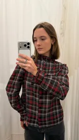 Camisa de algodón a cuadros con patrón en tonos rojo, negro y gris. Presenta cuello alto con volados, mangas largas con puños fruncidos y cierre frontal con botones.